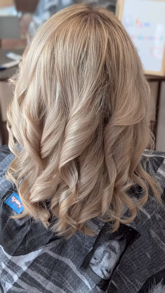 Efekt koloryzacji balayage w odcieniu ciepłego, świetlistego blondu na włosach ułożonych w eleganckie, grube loki.