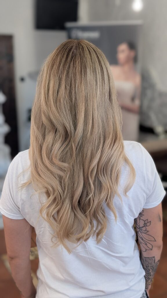 Widok od tyłu na długie, falowane włosy po koloryzacji balayage w chłodnych odcieniach blondu, wykonanej w salonie Colors Premium Słubice.